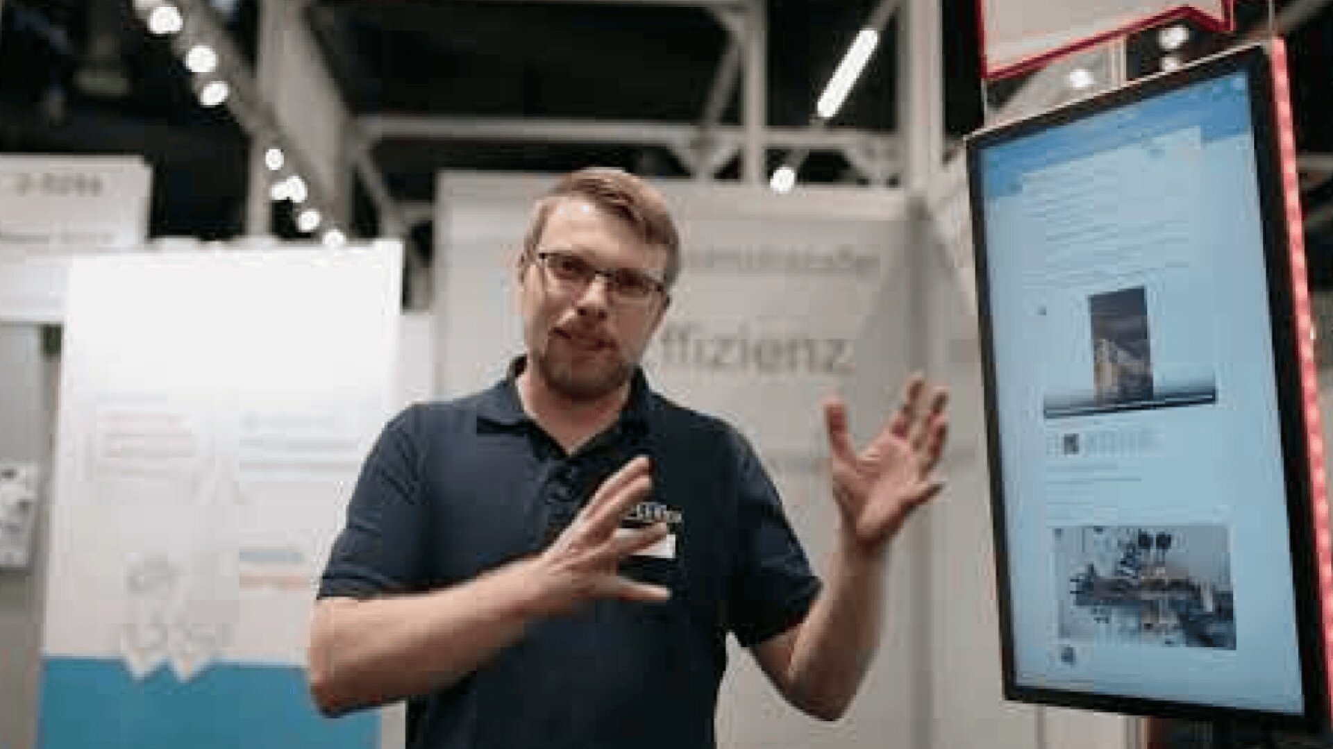 Ein Mann mit Brille und Bart gestikuliert, während er vor einem digitalen Bildschirm spricht, auf dem eine Website angezeigt wird. Es handelt sich offenbar um eine Technologie- oder Wirtschaftsmesse mit Informationsständen im Hintergrund.
