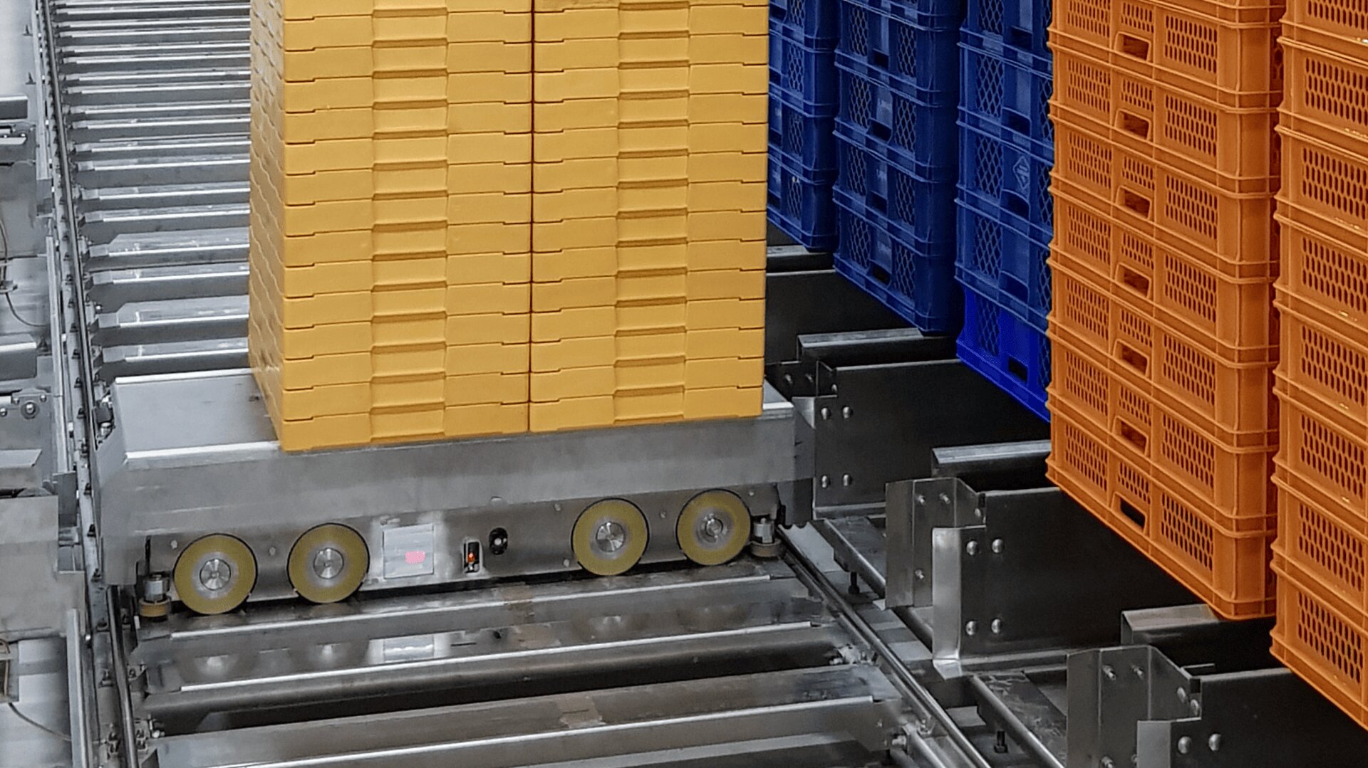 A robotic cart transports a tall stack of yellow plastic trays on a metal track system, with additional blue and orange trays stacked nearby in a warehouse or factory setting.