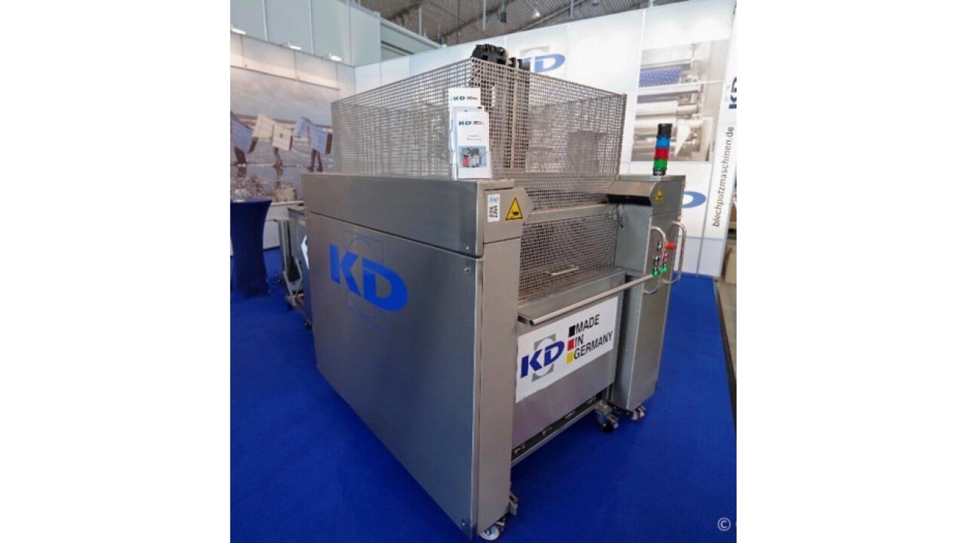 A large industrial machine with KD branding is displayed on a blue carpet at a trade show. The machine has a metal exterior, safety cages, control buttons, and a sign displaying its name and features.