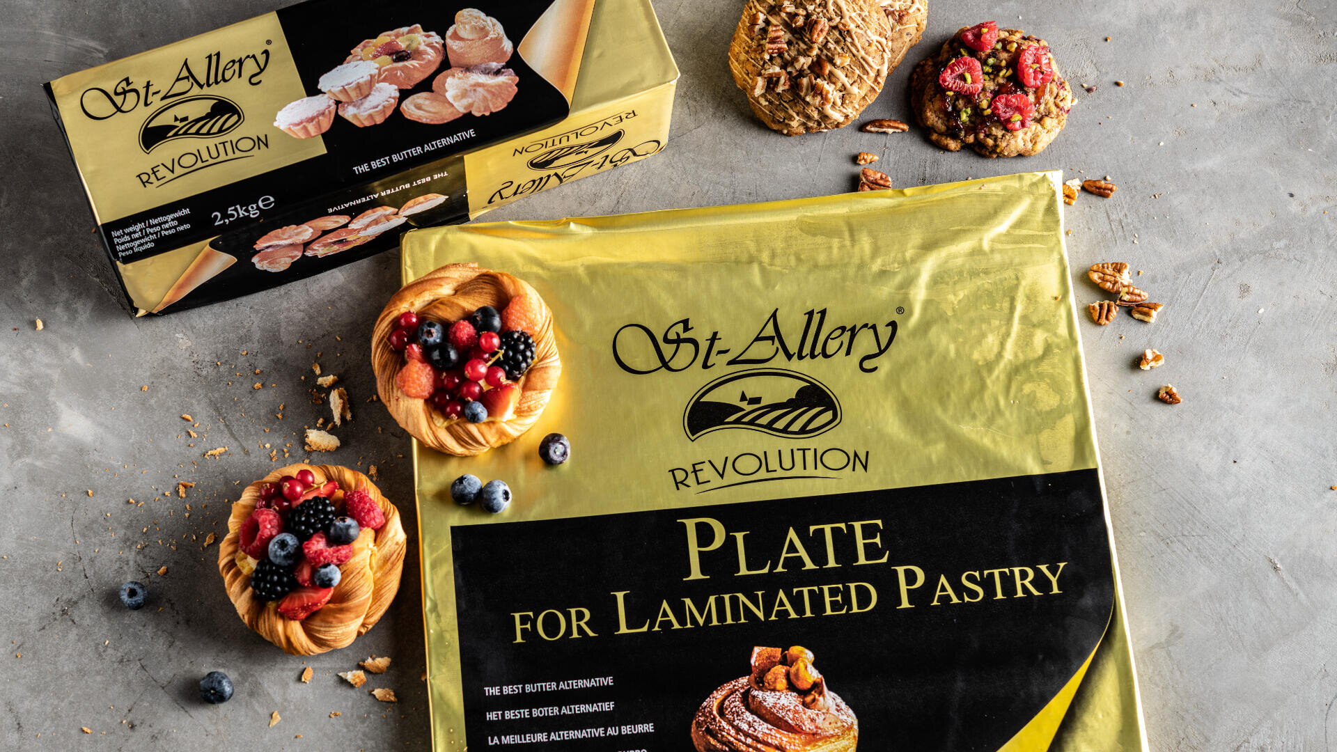A package of St. Allery Plate for Laminated Pastry on a gray surface, with a pastry box, two small fruit tarts, assorted cookies, blueberries, and pecans scattered around.
