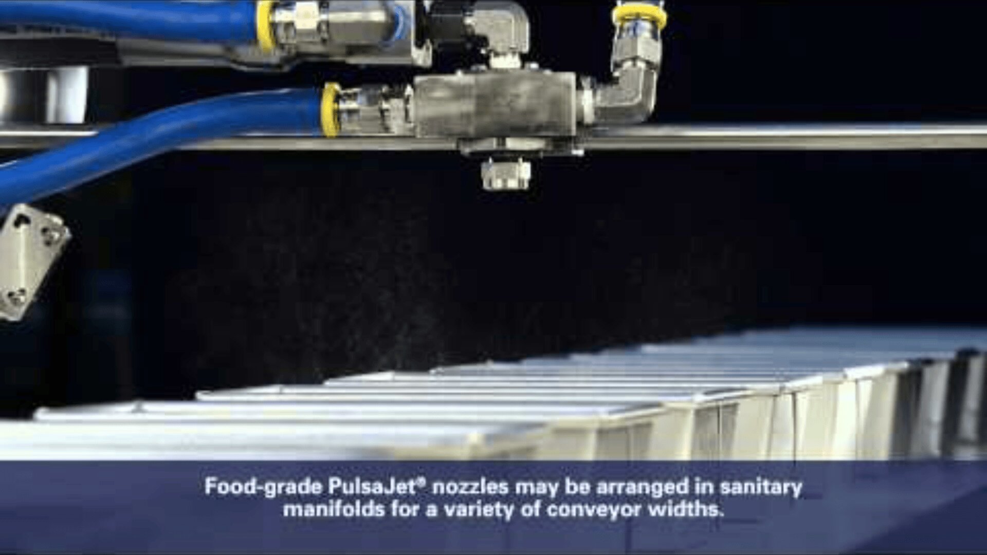 Nahaufnahme eines lebensmittelechten PulsaJet-Düsensystems, das Flüssigkeit über Behälter auf einem Förderband sprüht; über dem Förderband sind blaue Schläuche und Metallrohre sichtbar, mit einem Text, der die hygienischen Verteileranordnungen beschreibt.