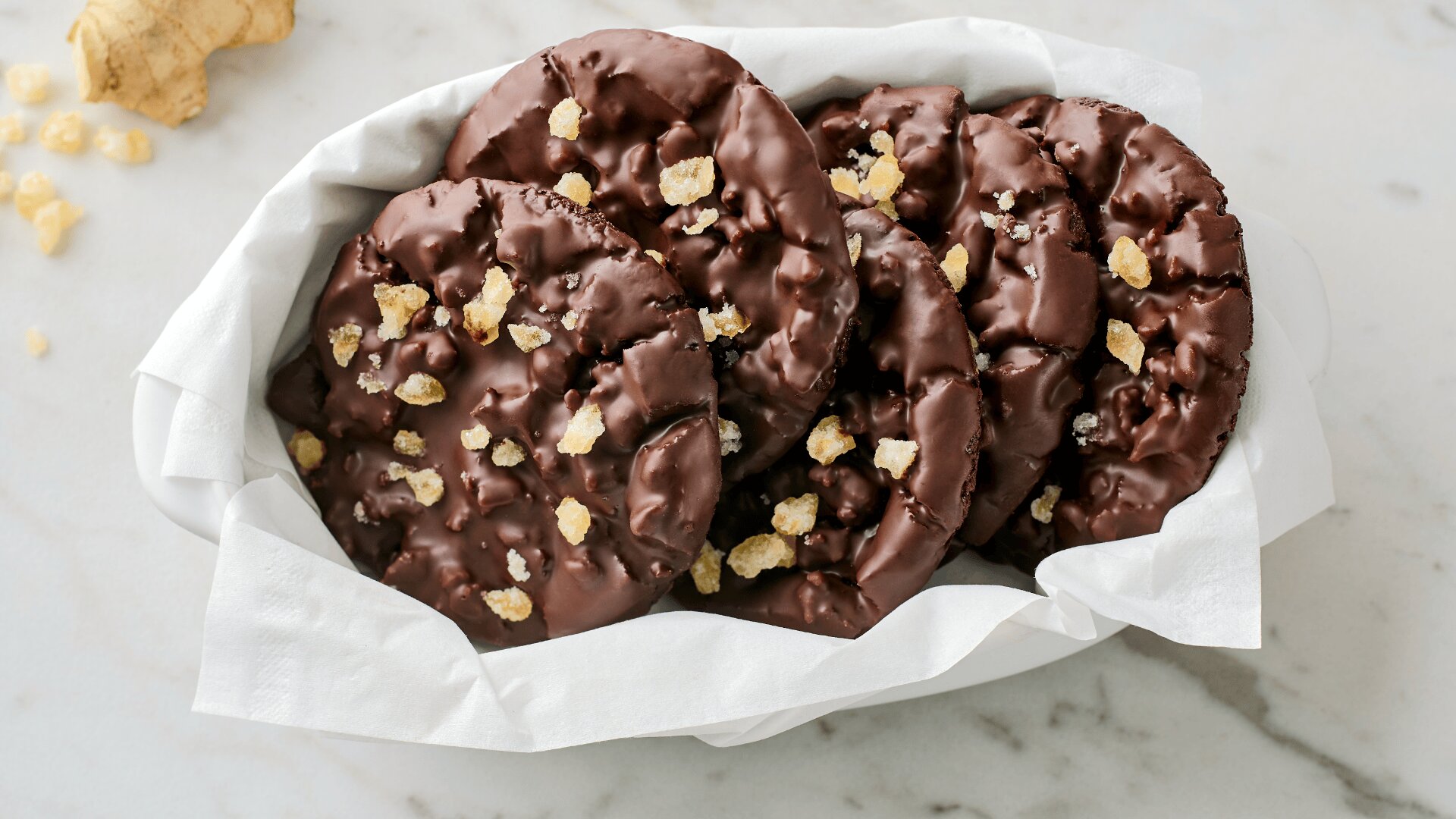 A white basket lined with parchment paper holds several chocolate cookies topped with pieces of candied ginger, placed on a light marble surface.