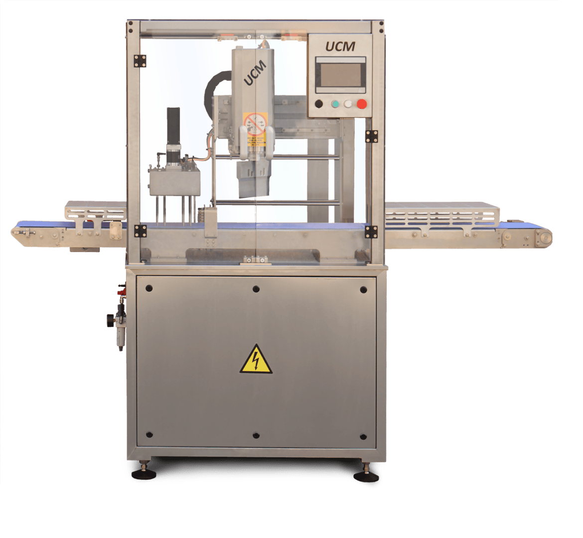 A stainless steel industrial machine with a transparent front panel, control screen, and conveyor belts on both sides. The machine has a caution sign for electricity on the lower panel and the label UCM at the top.