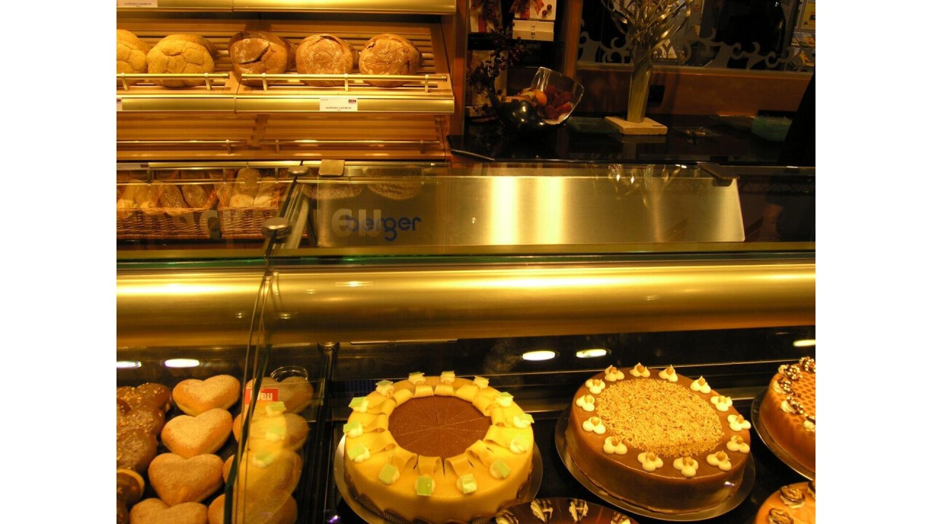 A bakery display case with assorted cakes and pastries, including heart-shaped cookies and round cakes with decorative toppings, and shelves of bread rolls in the background.
