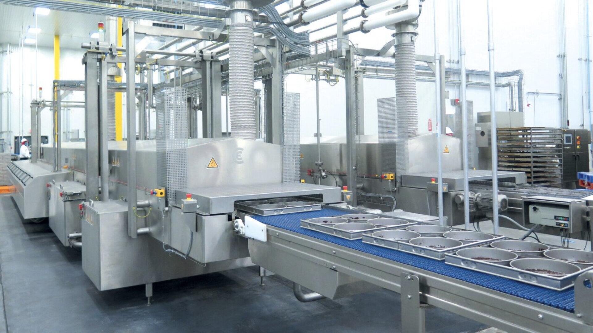A modern industrial food processing facility with stainless steel machines, conveyor belts, and trays, all inside a clean, well-lit production area with pipes and vents overhead.