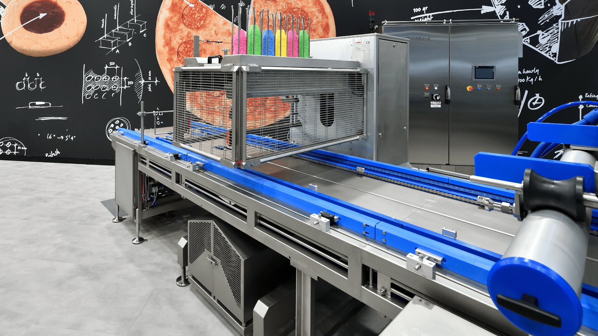 A modern industrial food processing machine with metal and blue components, colorful knives on top, and a large pizza image with white diagrams in the background.