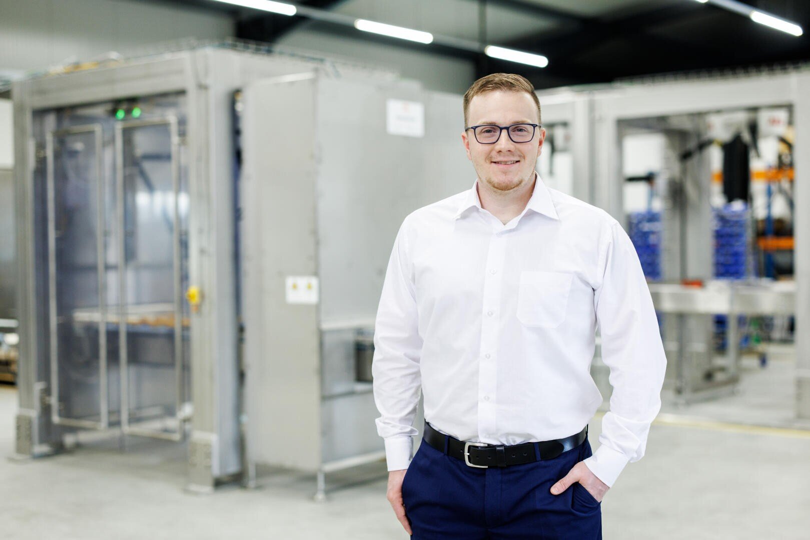 Ein Mann mit Brille und weißem Hemd steht lächelnd vor Industrieanlagen in einer hell erleuchteten Fabrik oder Lagerhalle.