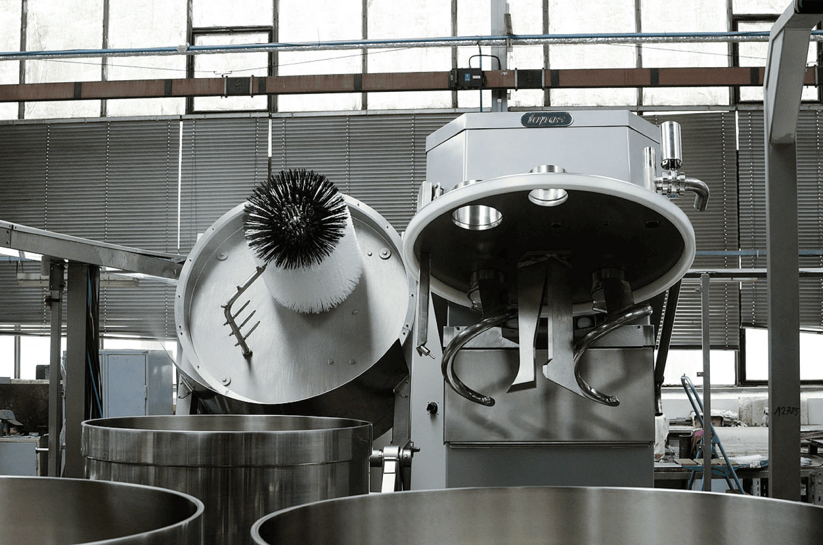 A large industrial food mixer with an open lid and visible mixing paddles stands in a factory setting. The lid has a circular brush attached, and stainless steel containers are positioned nearby.