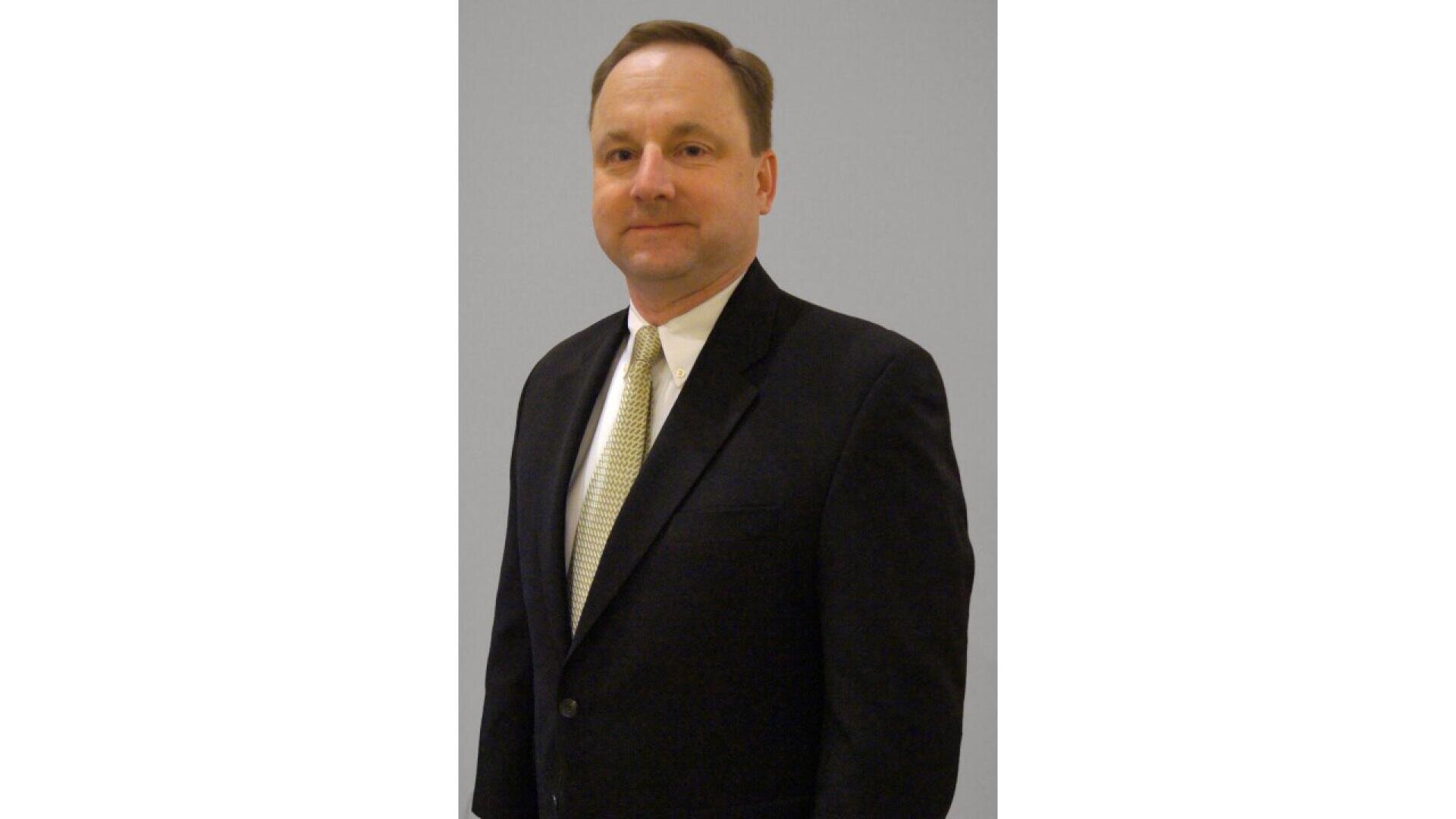 A man in a dark suit with a white dress shirt and light-colored tie stands against a plain, light gray background, facing slightly to the left and looking at the camera with a neutral expression.