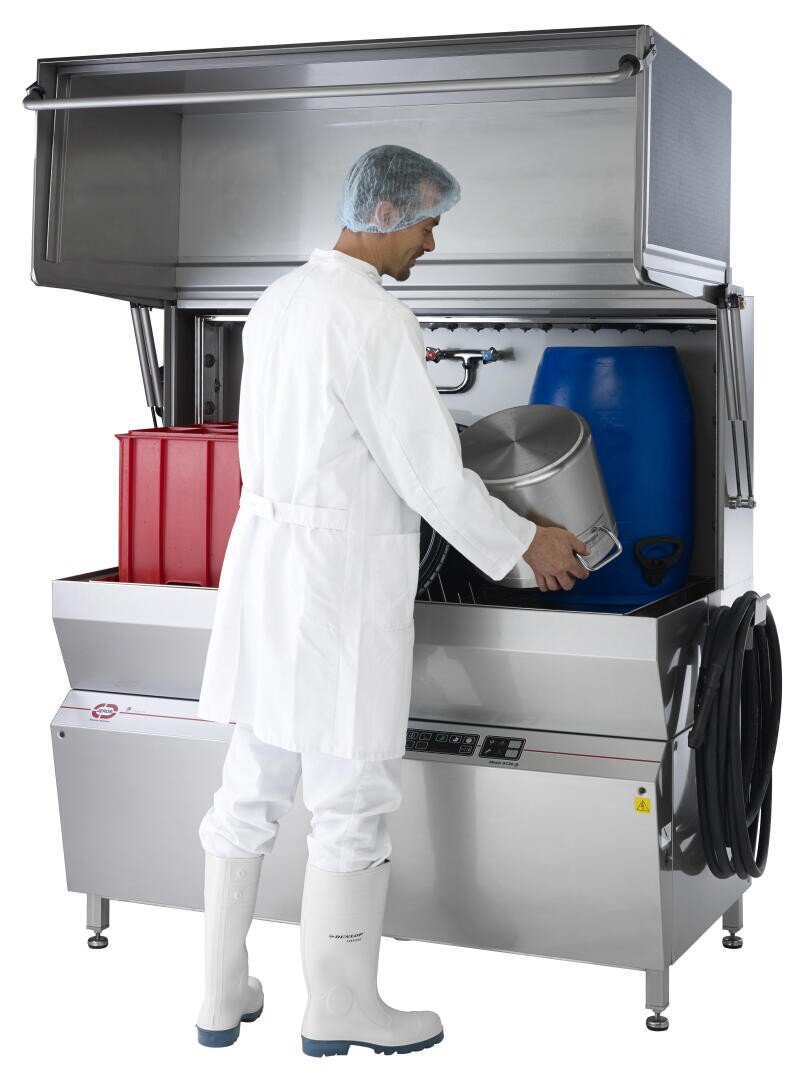 A person in white protective clothing and boots places a large metal container into an industrial washing machine with red and blue plastic bins inside. The machines lid is open, and various hoses are visible on the side.