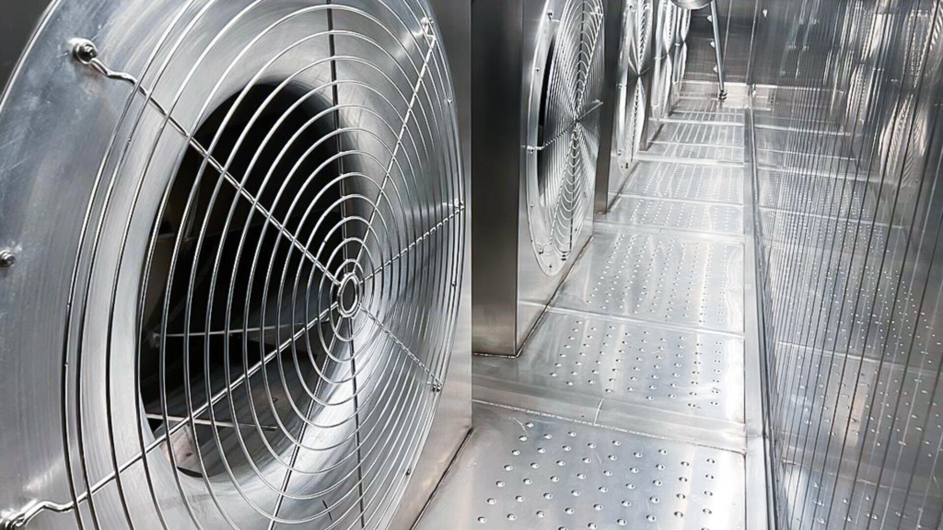 Large industrial metal fans and vented panels inside a shiny, metallic ventilation or air duct system, with a perspective showing both the fans and the long, reflective corridor.