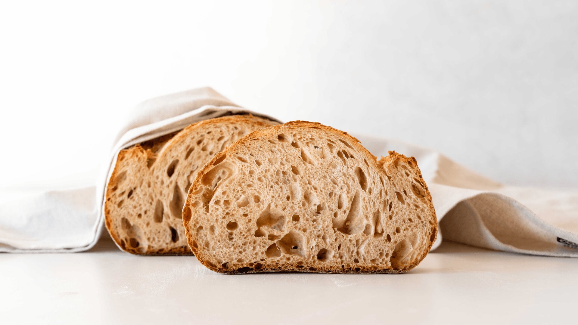 Zwei Scheiben rustikales Sauerteigbrot mit einer luftigen Krume werden auf eine weiße Fläche gelegt, die teilweise mit einem weißen Tuch vor einem hellen Hintergrund bedeckt ist.