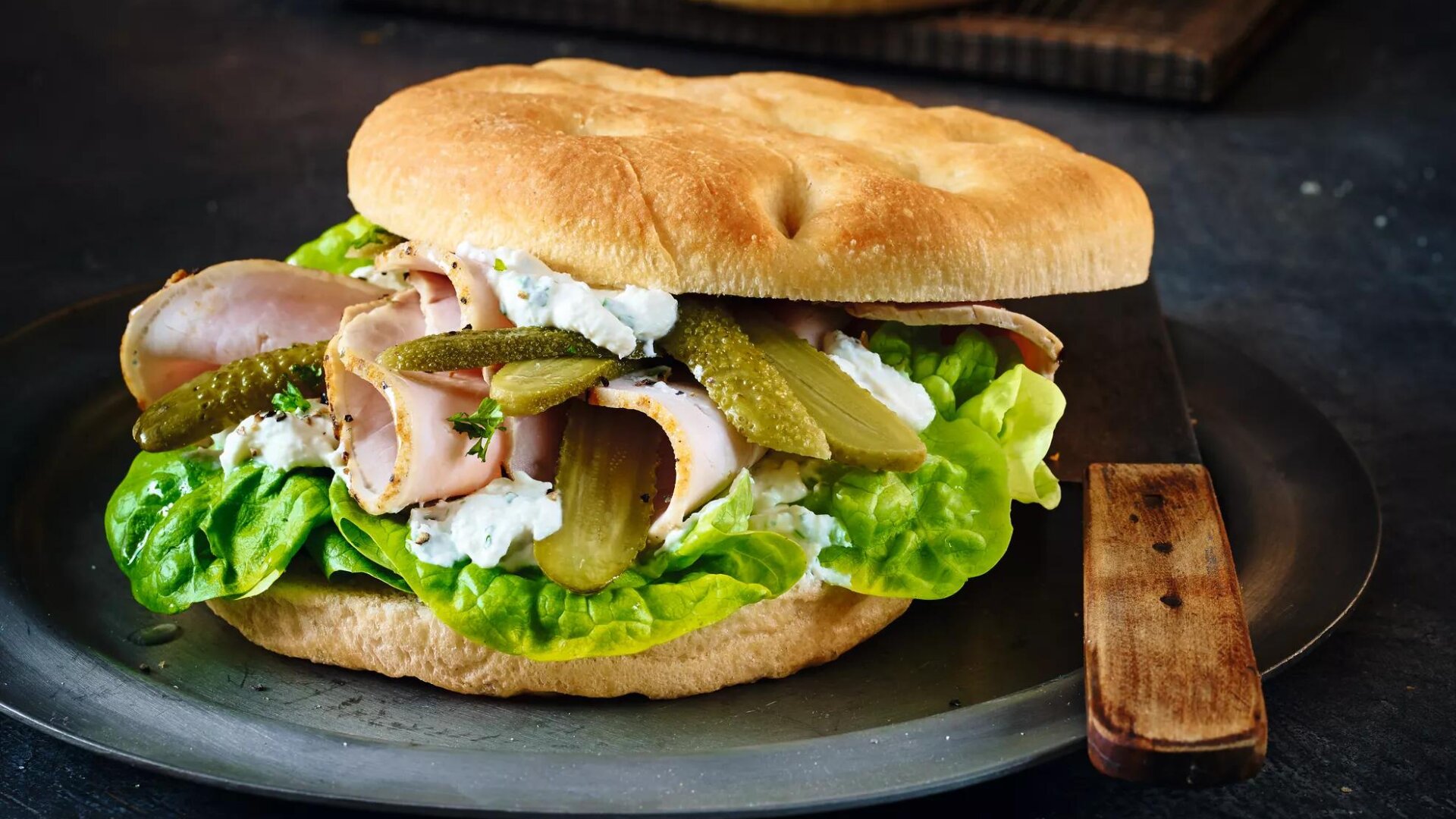 Ein Sandwich auf einem runden Fladenbrot, gefüllt mit Blattsalat, Puten- oder Schinkenscheiben, cremigem Weißkäseaufstrich und in Scheiben geschnittenen Essiggurken, serviert auf einem dunklen Teller mit einem Messer mit Holzgriff daneben.