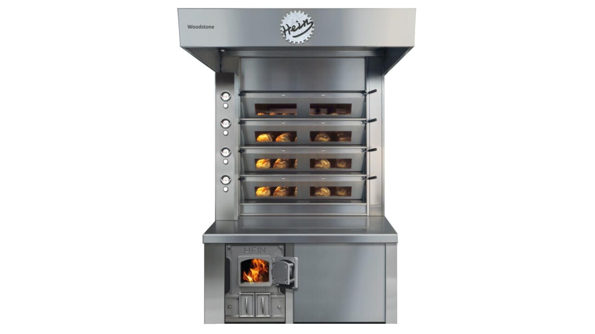 A commercial stainless steel bread oven with four baking chambers, each holding loaves of bread. Below the chambers is a wood-fired compartment with a visible flame behind a small door.