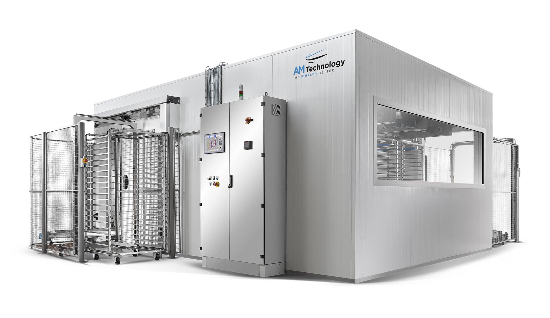 Large, modern industrial machine unit with control panel and metal racks, housed in a white enclosure labeled “AM Technology” with large windows and metal carts beside it.