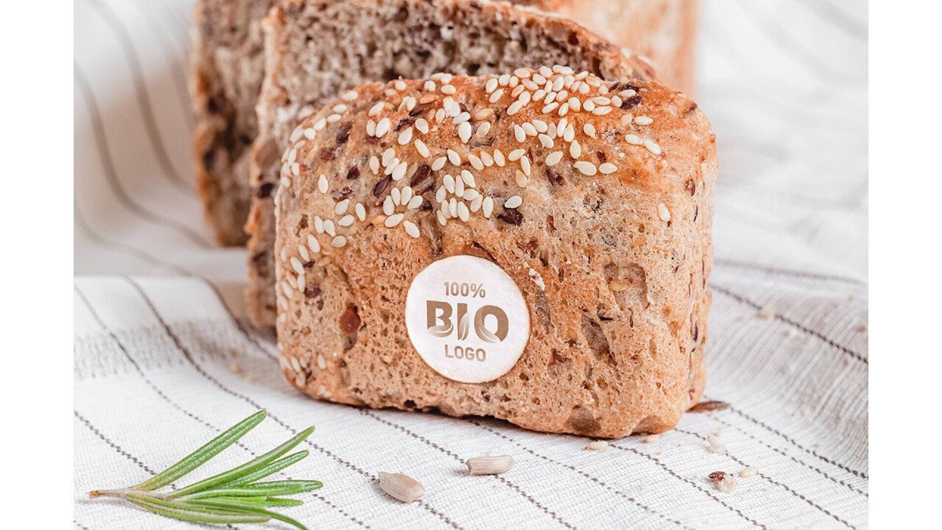 A loaf of multigrain bread topped with sesame seeds sits on a striped cloth. A round label on the bread reads 100% BIO LOGO. A sprig of rosemary and sunflower seeds are nearby.