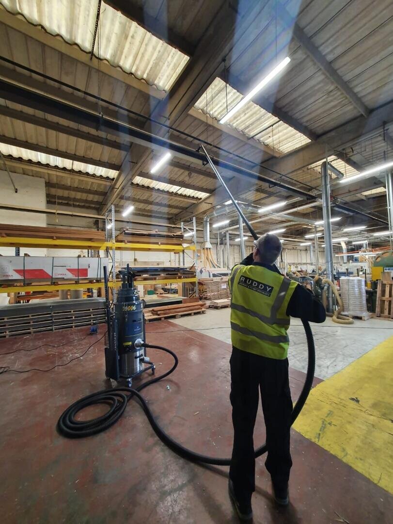 Ein Arbeiter in einer Warnweste reinigt mit einer langen Saugstange Rohre über der Decke in einer geräumigen Industriehalle. Im Hintergrund sind Stapel von Materialien und Geräten zu sehen.