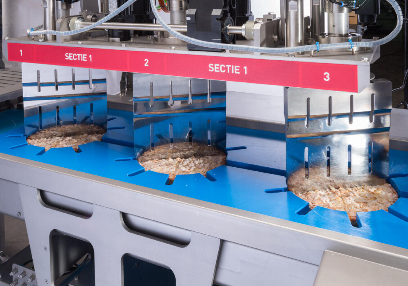 A food processing machine slices circular portions of a product on a blue conveyor under metal blades, labeled “SECTIE 1” above each section.