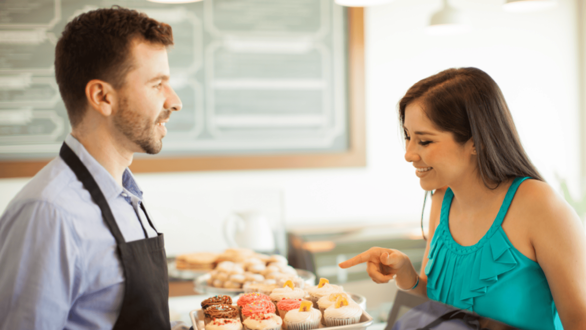 A man in an apron shows a tray of assorted cupcakes to a smiling woman in a teal top, who is pointing at a cupcake while standing at a bakery counter.