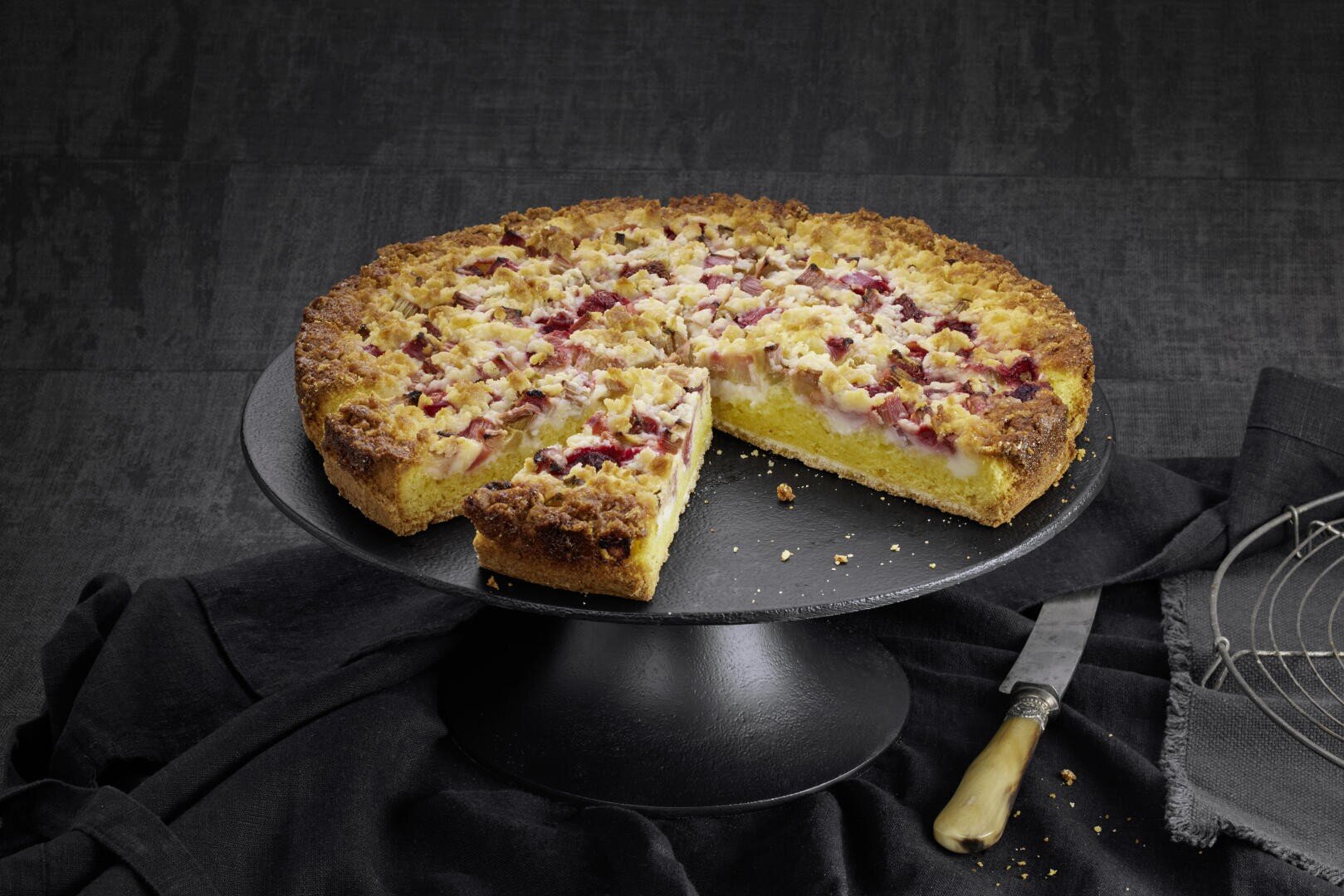 Ein runder, mit Krümeln belegter Kuchen, von dem eine Scheibe entfernt wurde, steht auf einem schwarzen Kuchenständer. Ein Messer und ein Kühlgestell sind neben dem Ständer auf einem dunklen Stoffhintergrund platziert.