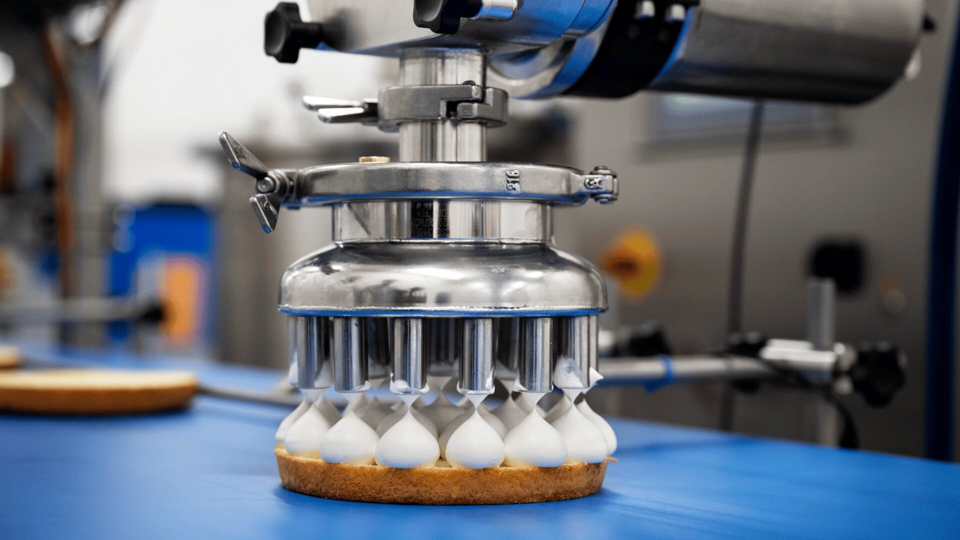 A close-up of a machine piping dollops of white cream onto a tart shell on a blue conveyor belt in a food production facility.