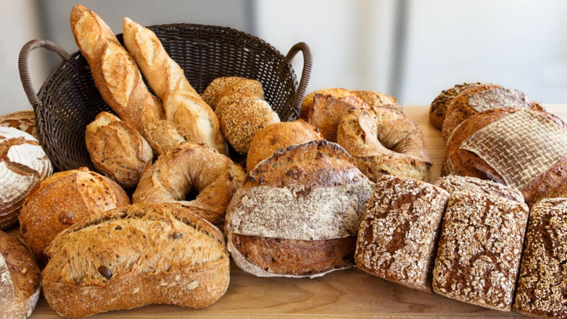 A variety of artisanal breads, including baguettes, round loaves, seed-covered rolls, and crusty rectangular loaves, are displayed on a wooden surface with a dark wicker basket in the background.