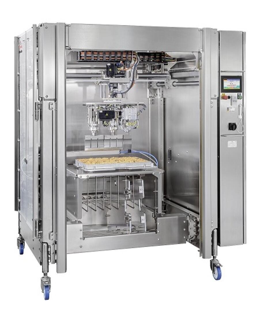 Eine große industrielle Lebensmittelverarbeitungsmaschine mit einem Metallrahmen, sichtbaren Kabeln und Komponenten und einem Touchscreen-Bedienfeld. Sie enthält ein Tablett mit Lebensmitteln und steht auf blauen Lenkrollen.