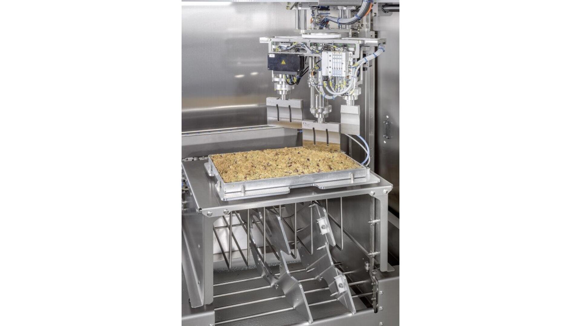 A food processing machine dispenses filling onto a rectangular tray of dough inside a stainless steel industrial environment, with wires and metal components visible.