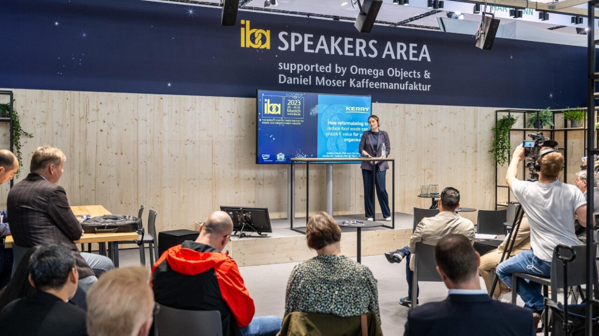 A speaker stands at a podium giving a presentation to a seated audience in a modern event space labeled “SPEAKERS AREA,” while photographers and attendees listen and take photos.