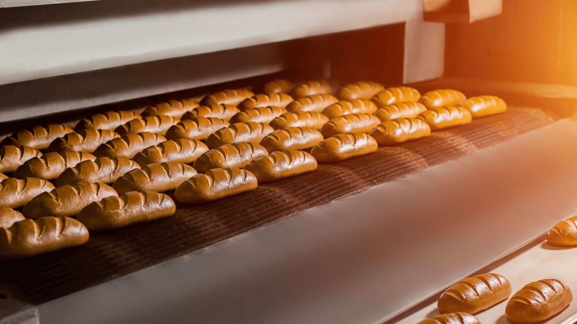Reihen von goldbraunen Brotlaiben, die auf einem Fließband in einem Industrieofen gebacken werden, wobei die warme Beleuchtung ihre frisch gebackenen Krusten hervorhebt.
