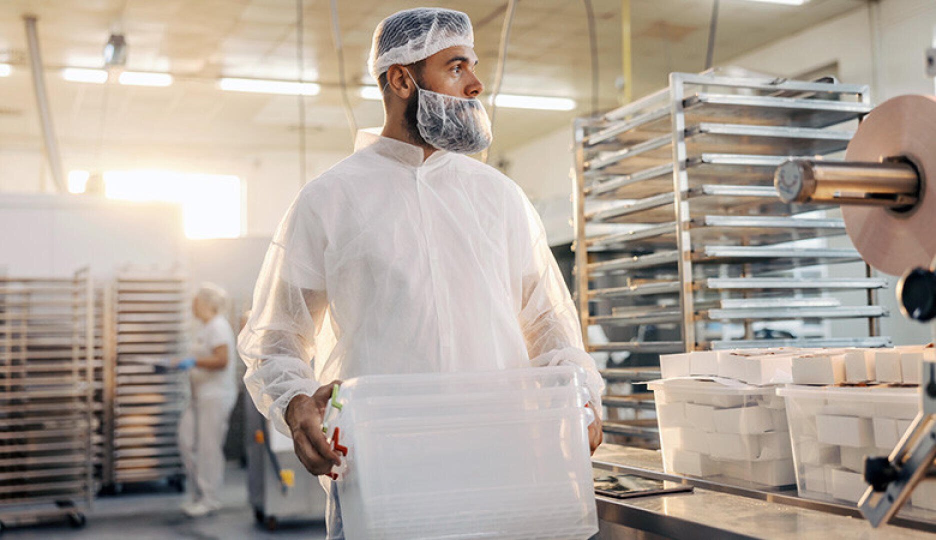 Ein Arbeiter mit Haarnetz, Bartschutz und weißem Schutzanzug hält einen durchsichtigen Plastikbehälter in einer Lebensmittelverarbeitungsanlage mit Metallgestellen und Tabletts im Hintergrund.
