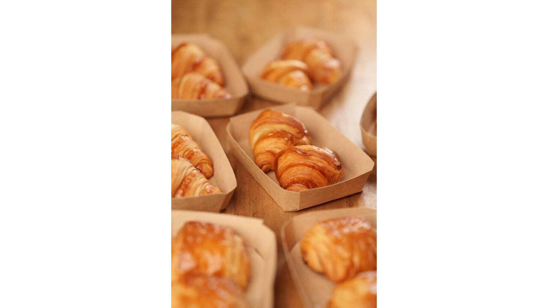 Mehrere goldbraune Croissants liegen in kleinen rechteckigen Papierschalen auf einer Holzplatte, bereit zum Servieren oder Verkaufen.