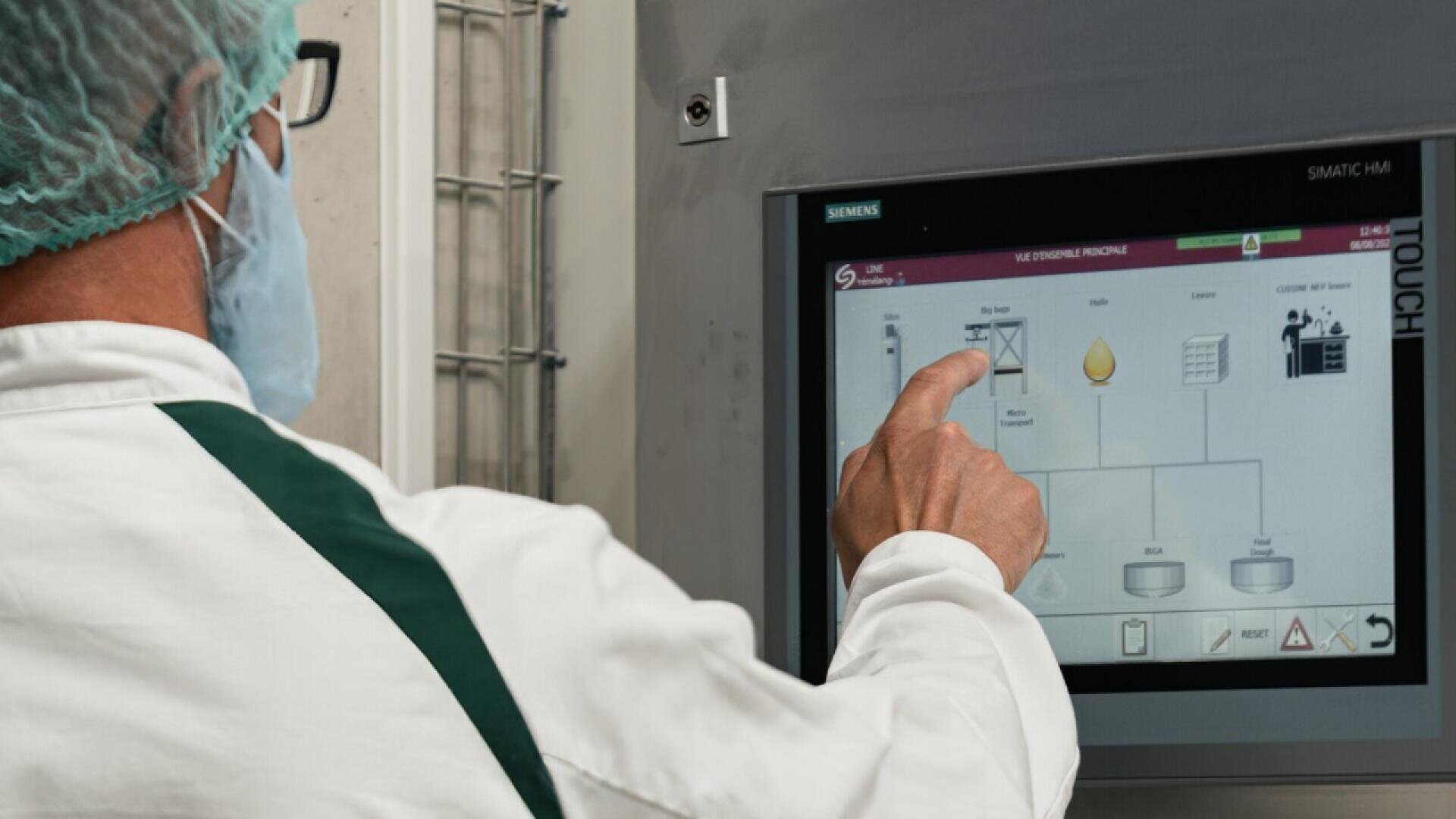 Eine Person mit Laborkittel, Haarnetz und Gesichtsmaske verwendet ein Touchscreen-Bedienfeld, auf dem ein industrielles Prozessdiagramm angezeigt wird.