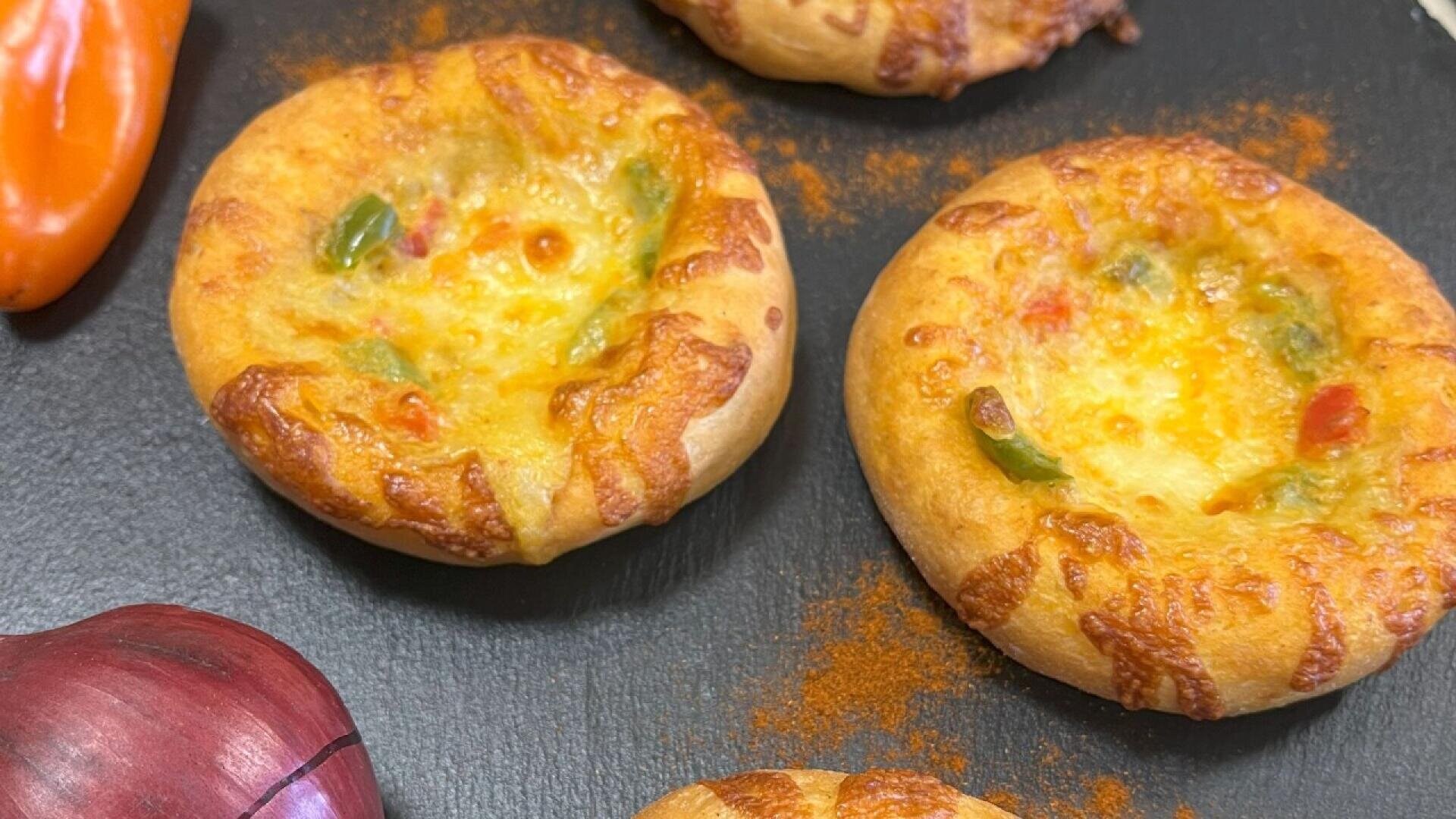Three mini round bread pizzas with melted cheese and colorful bell peppers on a black surface, next to a piece of red onion and an orange pepper.