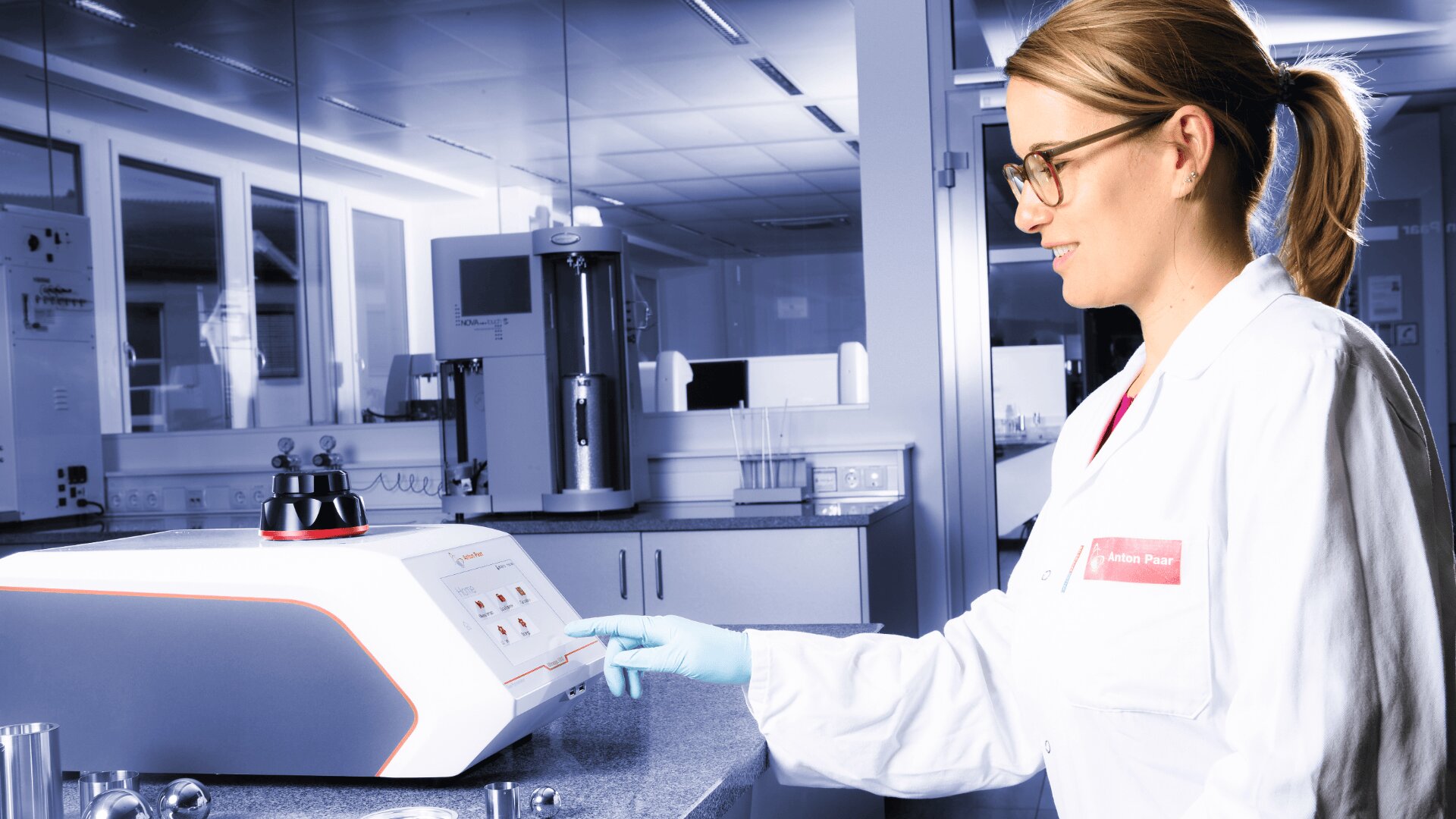A woman in a white lab coat and safety glasses operates a scientific instrument in a modern laboratory setting. She is pressing a button on the device, standing at a counter with lab equipment.