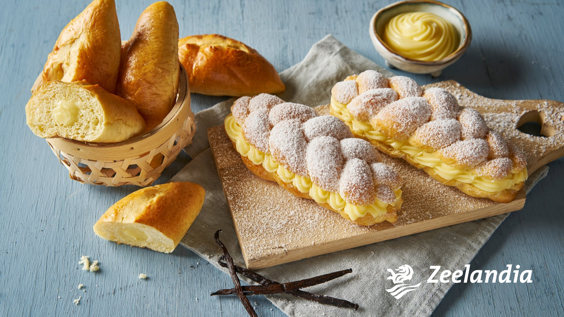 Lange Eclairs und gefüllte Brötchen mit cremiger Vanillefüllung liegen auf einer Holzfläche, eines davon auf einem mit Puderzucker bestäubten Schneidebrett. Zu sehen sind eine Schüssel Sahne, Vanilleschoten und das Zeelandia-Logo.