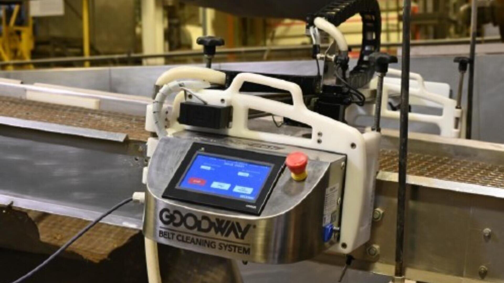 A Goodway belt cleaning system with a digital touchscreen and emergency stop button is mounted next to a conveyor belt in an industrial setting.