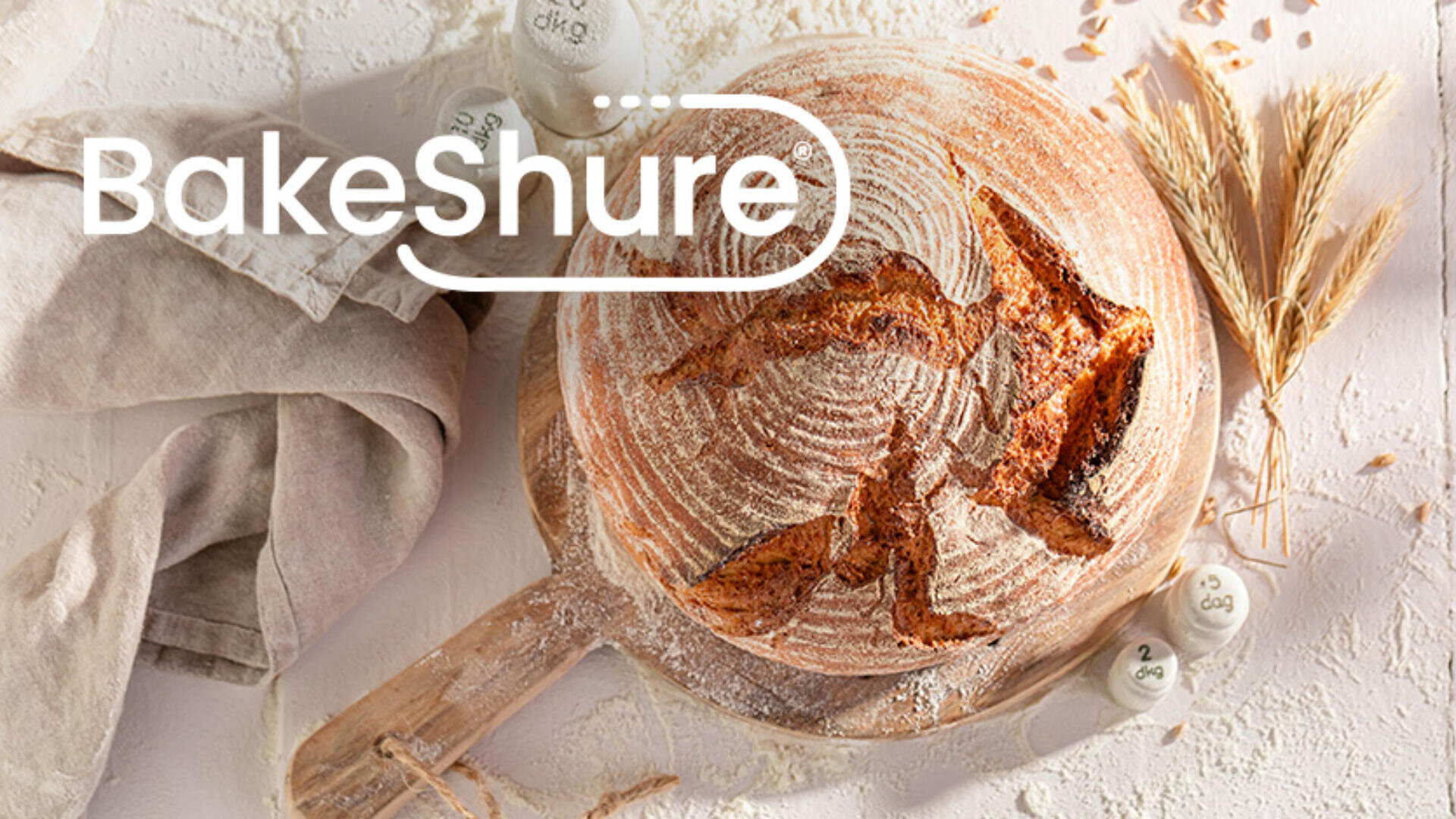 Round rustic loaf of bread on a wooden board with flour, a beige cloth, wheat stalks, and measuring spoons. The word BakeShure is overlaid in bold white text at the top left.