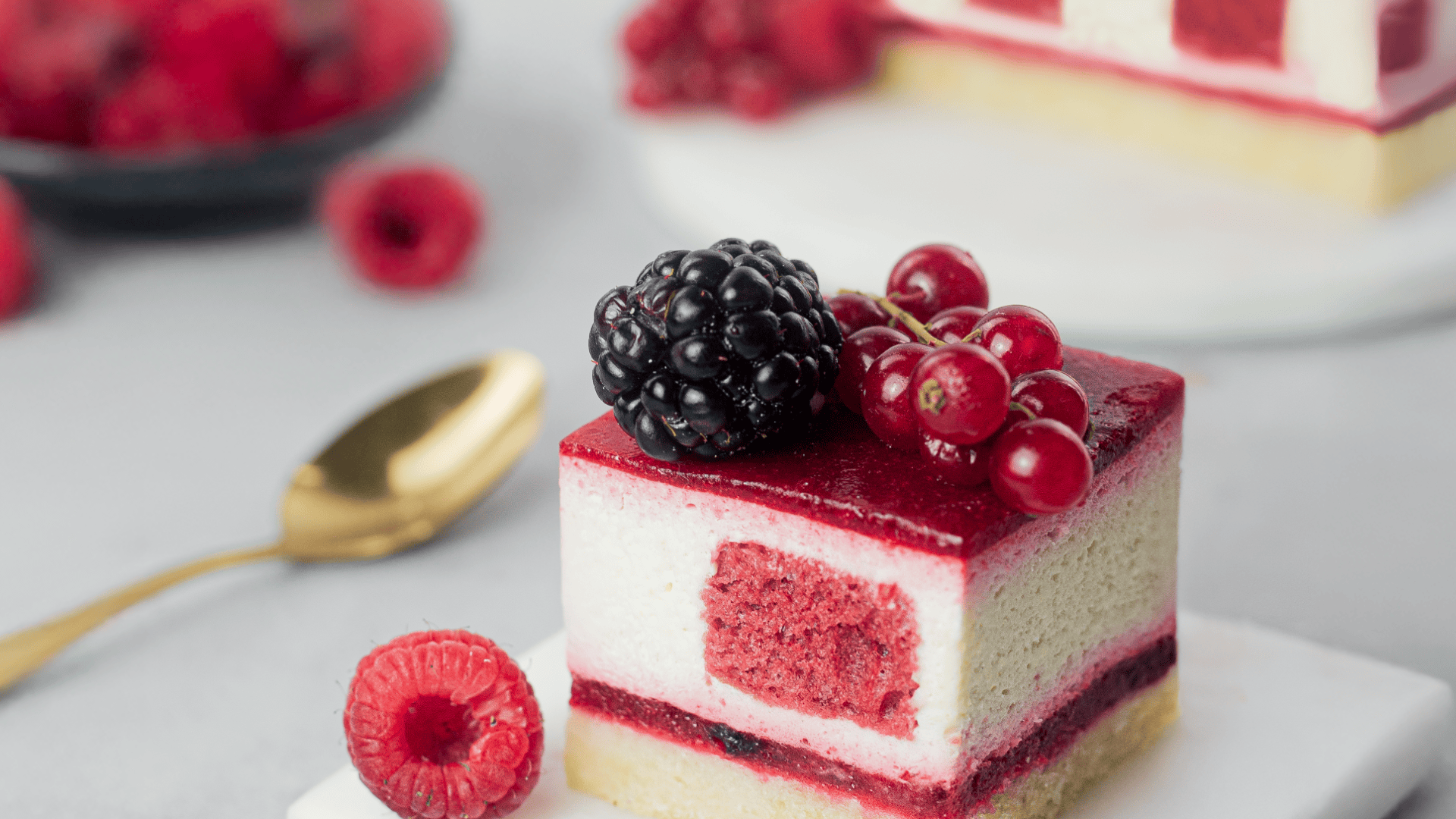A slice of layered berry mousse cake topped with a blackberry and red currants, garnished with a raspberry on the side. A gold spoon and more berries are blurred in the background.