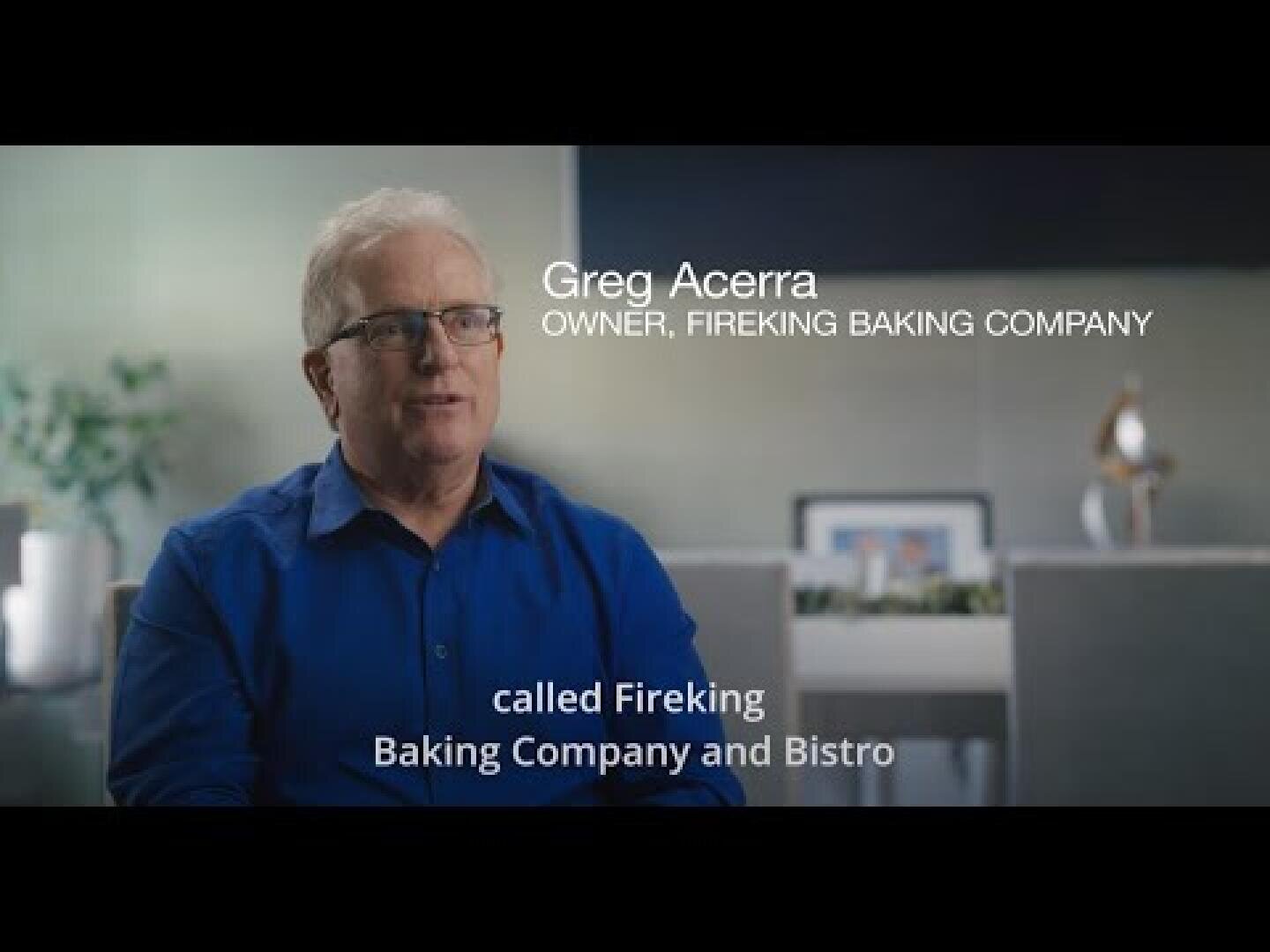 Ein Mann in einem blauen Hemd sitzt in einem Haus, auf dem Bild ist zu lesen: Greg Acerra, Inhaber, Fireking Baking Company, und darunter: Fireking Baking Company and Bistro. Der Hintergrund ist sanft verschwommen.