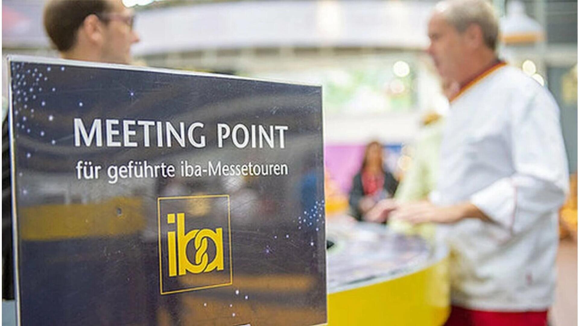 A close-up of a sign reading MEETING POINT für geführte iba-Messetouren with two people conversing in the blurred background at an indoor event or exhibition.