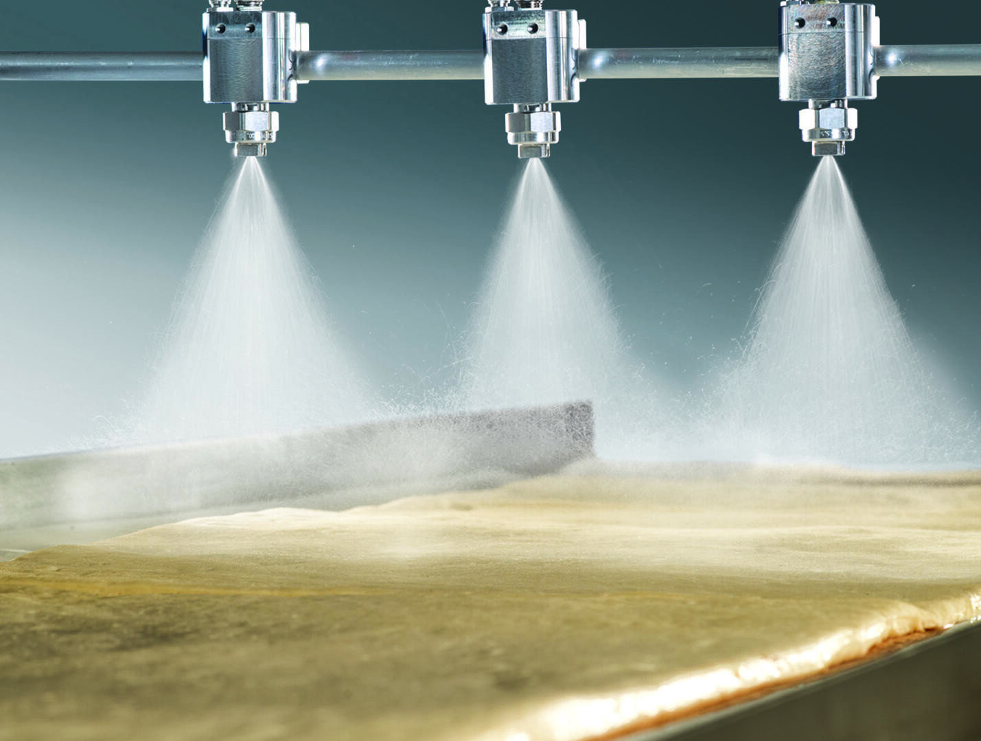 Drei Metalldüsen sprühen einen feinen Nebel über ein flaches, goldenes Materialblatt auf einem Förderband, wahrscheinlich als Teil eines industriellen Herstellungs- oder Lebensmittelverarbeitungsprozesses.
