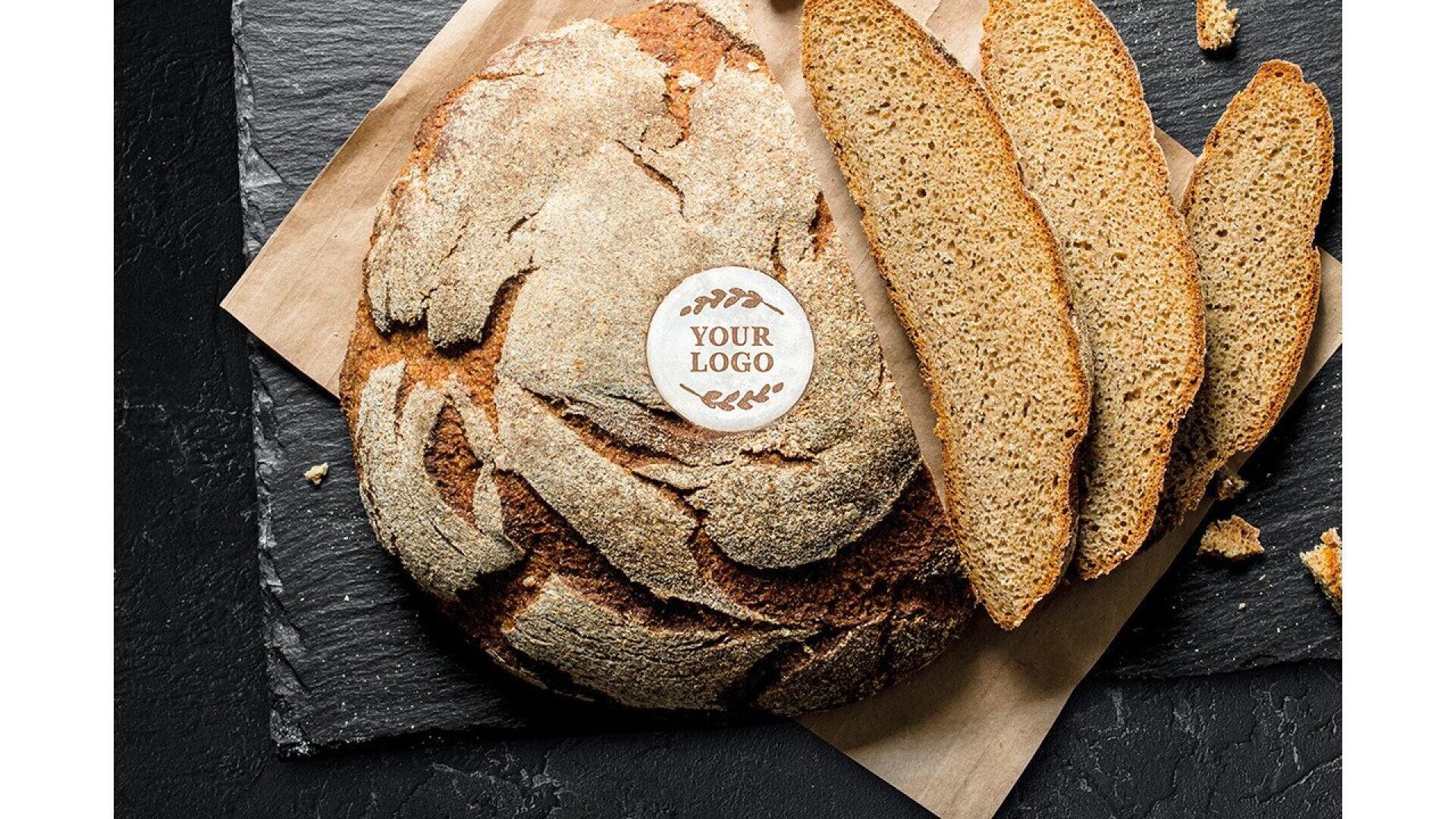 A round loaf of rustic bread with a Your Logo edible topper, partially sliced, sits on brown parchment atop a dark slate surface. Three slices are fanned out next to the unsliced portion.
