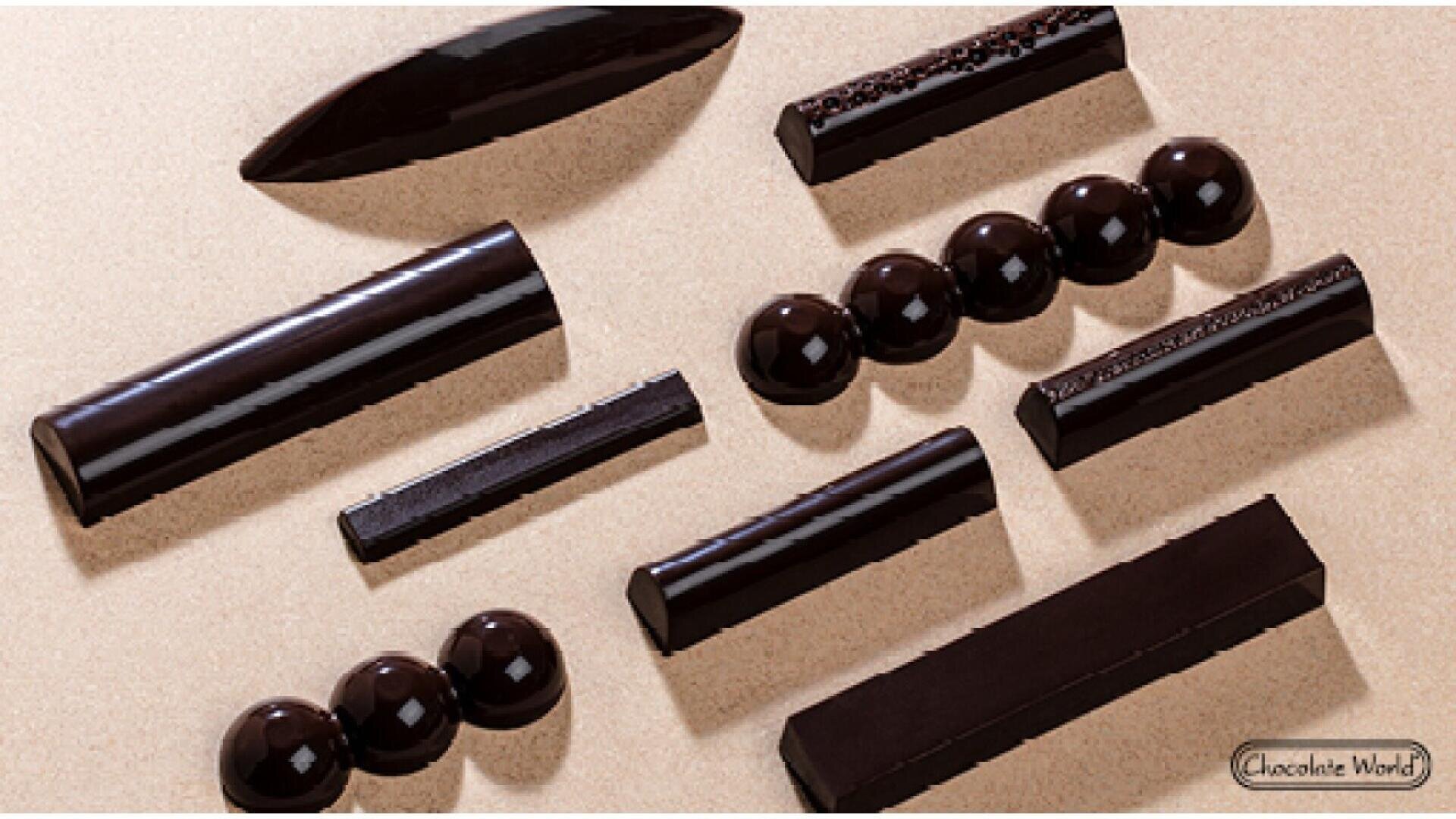 Verschiedene glänzende dunkle Schokoladenstücke in unterschiedlichen Formen - Kugeln, Rechtecke und Tafeln - sind auf einer beigen Oberfläche arrangiert. Die Pralinen haben eine glatte, glänzende Oberfläche, und einige weisen deutliche Texturen oder Muster auf.