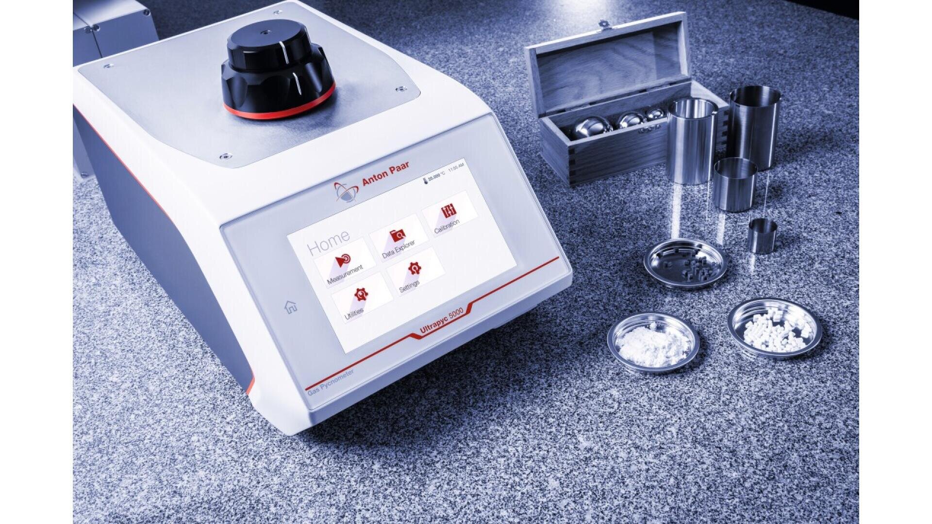 Auf einer Arbeitsplatte steht ein Laborgerät mit Touchscreen-Oberfläche neben verschiedenen Metallbehältern, Pulvern und einer Holzkiste mit Metallkugeln, möglicherweise zum Mahlen oder Zerkleinern von Proben.