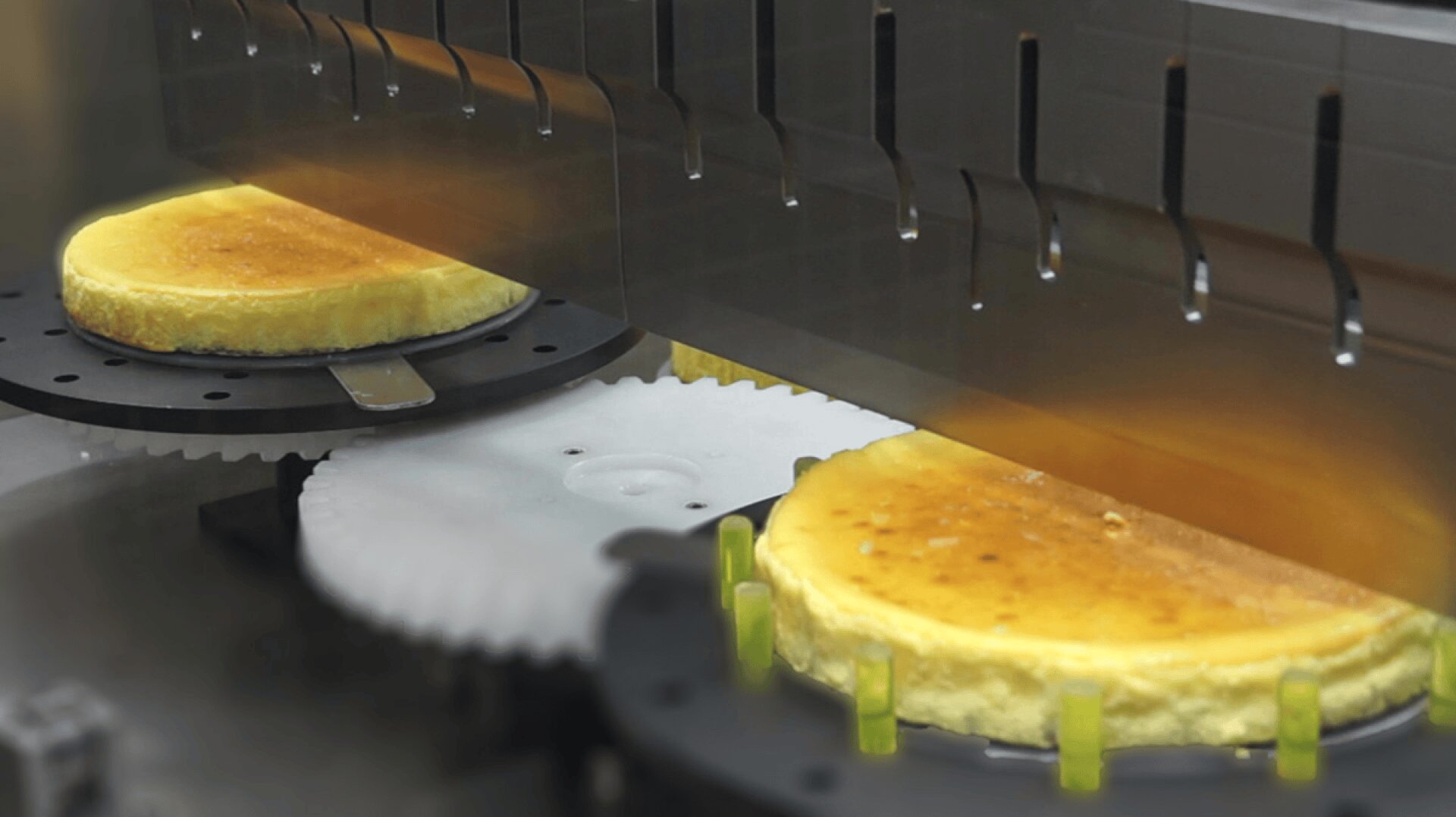 Two cheesecakes move along a conveyor in an automated factory line, passing beneath a machine equipped with nozzles, likely for applying toppings or glazes.