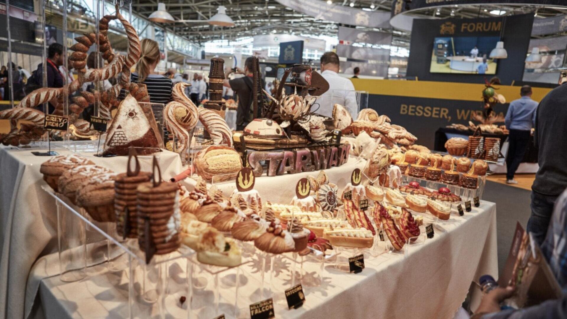 A large bakery display features a variety of breads, pastries, and cakes arranged on tables at a food exhibition. Shoppers and exhibitors walk in the background among booths and displays.