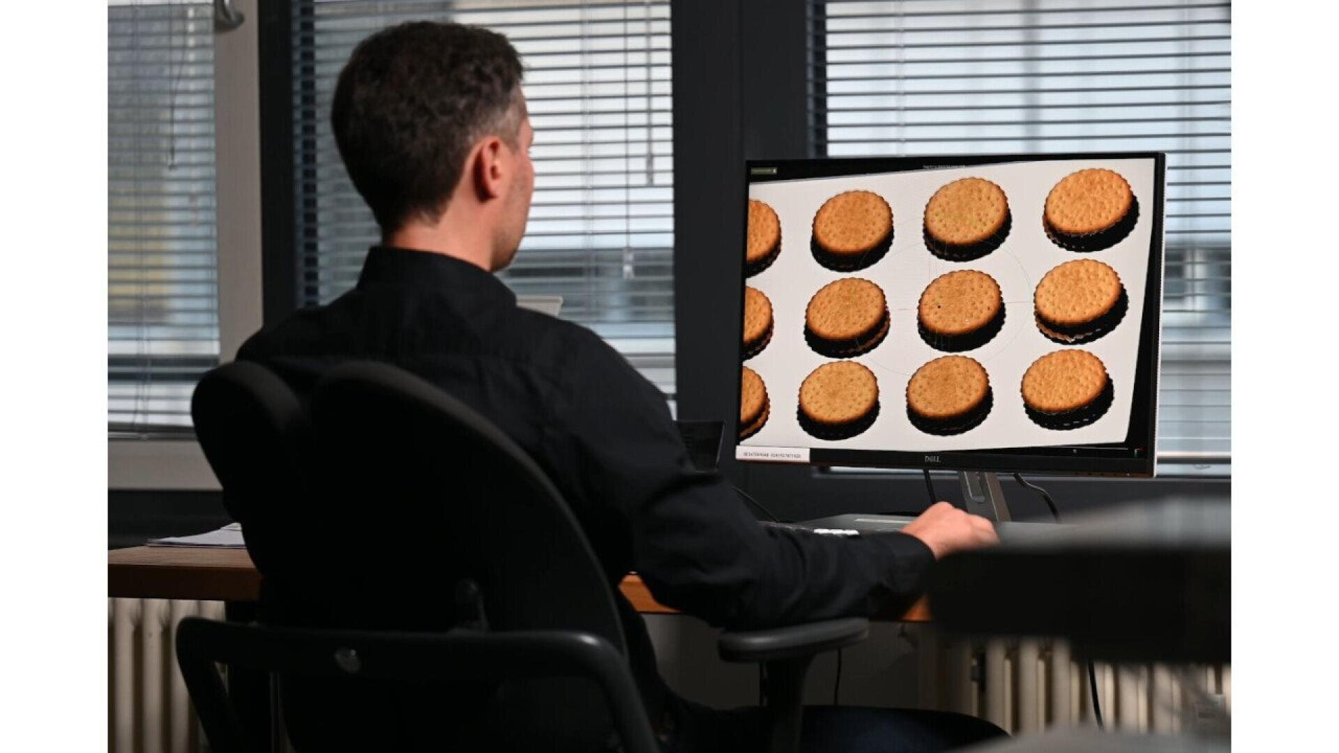 A man sitting at a desk looks at a computer monitor displaying multiple images of round cookies or biscuits on a white background.