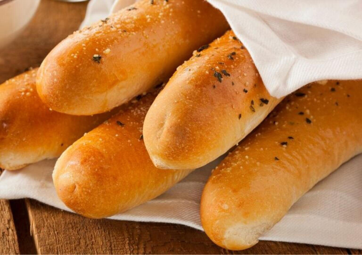 Four golden-brown breadsticks sprinkled with herbs and salt are partially wrapped in a white cloth, resting on a wooden surface.