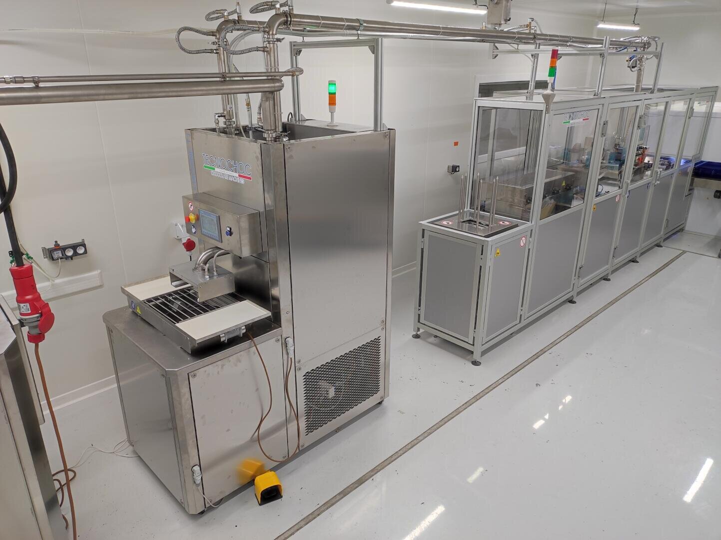 A clean, bright laboratory with industrial stainless steel equipment, control panels, indicator lights, and enclosed machinery along the wall, used for automated manufacturing or processing. The floor is shiny and spotless.